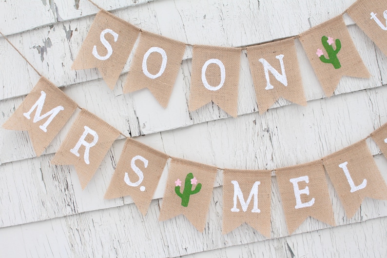 May include: A burlap banner with the words "Soon to be Mrs." and a cactus design. The banner is strung on a white wooden wall.