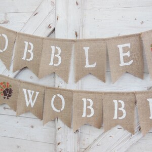 May include: A burlap banner with white stenciled letters spelling "GOBBLE YA WOBBL" and a turkey graphic. The banner is made of natural-colored burlap and is suitable for Thanksgiving or fall decor.