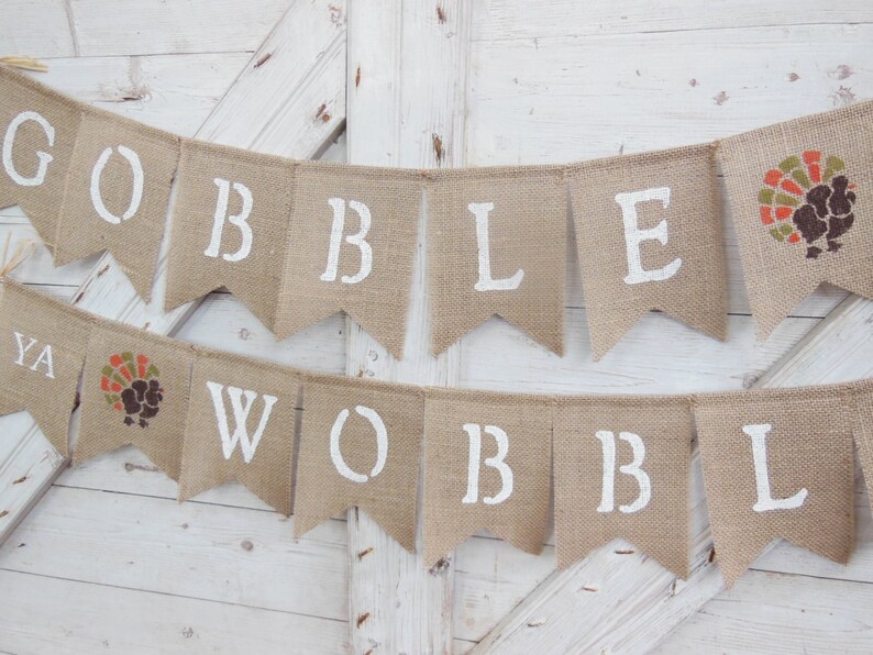 May include: A burlap banner with white stenciled letters spelling "GOBBLE YA WOBBL" and a turkey graphic. The banner is made of natural-colored burlap and is suitable for Thanksgiving or fall decor.