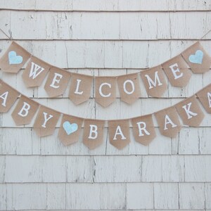 Puede incluir: Una pancarta de arpillera con las palabras "Welcome Baby Barakat" en letras blancas. La pancarta est&aacute; decorada con corazones azules.