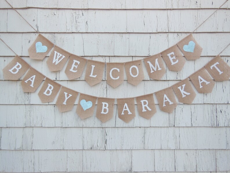 Puede incluir: Una pancarta de arpillera con las palabras "Welcome Baby Barakat" en letras blancas. La pancarta est&aacute; decorada con corazones azules.