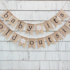 May include: A burlap banner with the text "Baby it's cold outside" in black lettering. The banner has snowflakes on two of the flags.