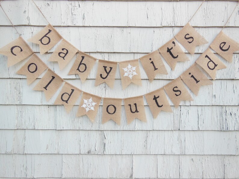 May include: A burlap banner with the text "Baby it's cold outside" in black lettering. The banner has snowflakes on two of the flags.