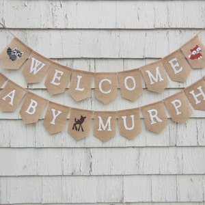 Puede incluir: Una pancarta de arpillera con las palabras "Welcome Baby Murphy" en letras blancas. La pancarta est&aacute; decorada con peque&ntilde;as ilustraciones de animales, incluyendo un mapache, un zorro y un ciervo.