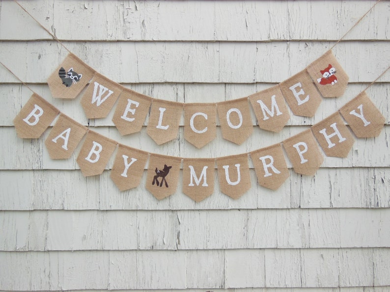 Puede incluir: Una pancarta de arpillera con las palabras "Welcome Baby Murphy" en letras blancas. La pancarta est&aacute; decorada con peque&ntilde;as ilustraciones de animales, incluyendo un mapache, un zorro y un ciervo.