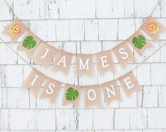 May include: A burlap banner with the words "JAMES IS ONE" in white letters. The banner is decorated with green leaves and small lion illustrations.