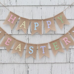 May include: A burlap banner with the words "Happy Easter" in pastel colors. The banner is strung on a white wooden wall.