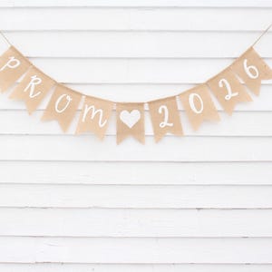 May include: A banner made of tan burlap flags with the words "PROM 2016" in white script, strung on twine. A white heart is between the words "PROM" and "2016". The banner hangs against a white, horizontal-planked wall.