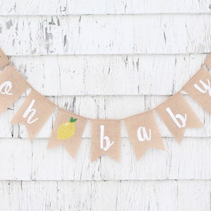 Peut inclure: Une banni&egrave;re en toile de jute avec les mots "oh baby" peints en blanc. Chaque lettre est sur un drapeau s&eacute;par&eacute; avec une illustration de citron jaune. La banni&egrave;re est accroch&eacute;e &agrave; un mur en bois blanc.