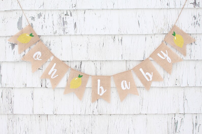 Peut inclure: Une banni&egrave;re en toile de jute avec les mots "oh baby" peints en blanc. Chaque lettre est sur un drapeau s&eacute;par&eacute; avec une illustration de citron jaune. La banni&egrave;re est accroch&eacute;e &agrave; un mur en bois blanc.