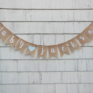 May include: A burlap banner with the words "baby Jackson" in white paint. A blue heart is between the words "baby" and "Jackson".