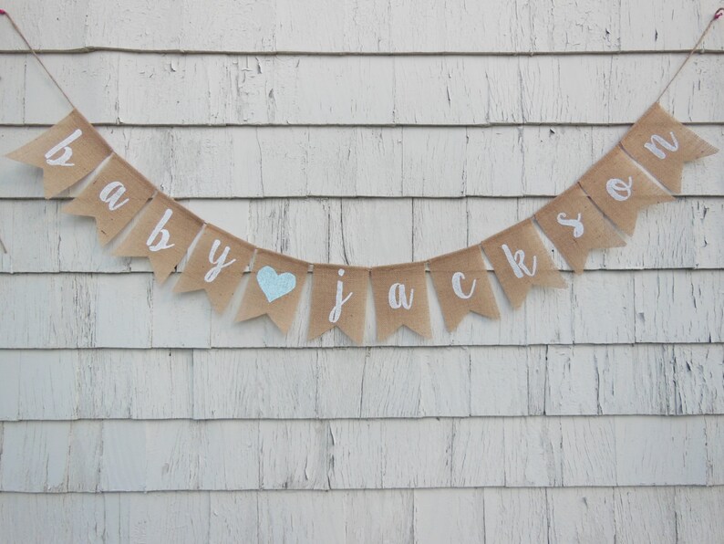 May include: A burlap banner with the words "baby Jackson" in white paint. A blue heart is between the words "baby" and "Jackson".