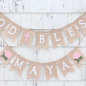 Puede incluir: Una pancarta de arpillera con las palabras "God Bless Maya" pintadas en blanco. La pancarta est&aacute; decorada con flores rosas y una cruz rosa.