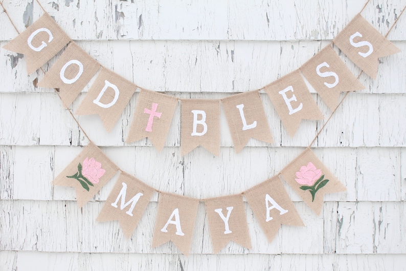 Puede incluir: Una pancarta de arpillera con las palabras "God Bless Maya" pintadas en blanco. La pancarta est&aacute; decorada con flores rosas y una cruz rosa.