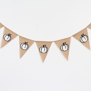May include: A burlap bunting featuring triangular flags, each printed with a black and white pumpkin design. The banner is strung on twine, ideal for autumn decorations. The pumpkins are centered on each flag.