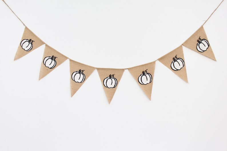 May include: A burlap bunting featuring triangular flags, each printed with a black and white pumpkin design. The banner is strung on twine, ideal for autumn decorations. The pumpkins are centered on each flag.