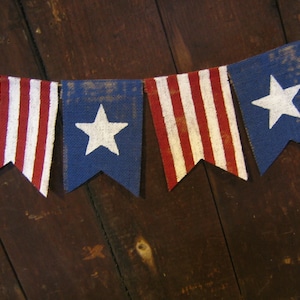 May include: A bunting banner with red, white, and blue fabric flags. The flags have a star design and stripes. The banner is hanging on a wooden surface.