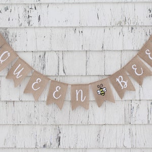May include: A burlap banner with the words "Queen Bee" in white lettering. The banner is decorated with small bee illustrations.