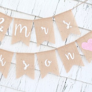 K&ouml;nnte beinhalten: Ein Banner aus Jute mit den Worten "Mr. & Mrs." und einem rosa Herz. Das Banner ist an einem wei&szlig;en Holzgrund befestigt.