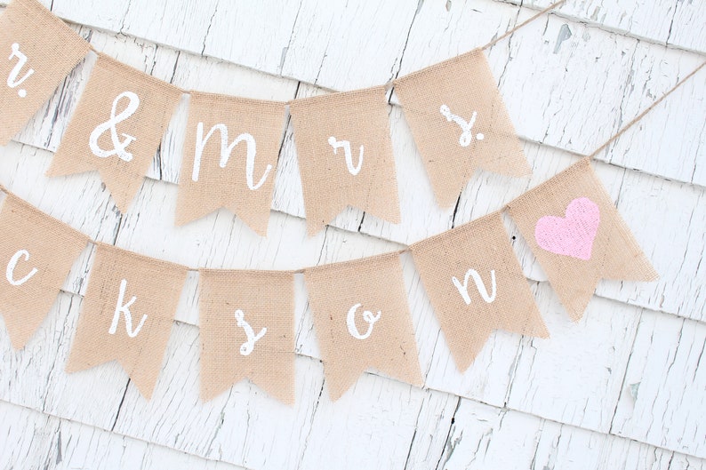 K&ouml;nnte beinhalten: Ein Banner aus Jute mit den Worten "Mr. & Mrs." und einem rosa Herz. Das Banner ist an einem wei&szlig;en Holzgrund befestigt.