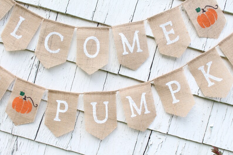 May include: A burlap banner with the words "Welcome Pumpkin" in white paint. The banner is decorated with small orange pumpkins.