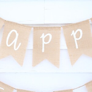 May include: A burlap banner with the word "HAPPY" spelled out in white letters. The banner is made of individual pennants strung together with twine. The background is a white wooden surface.