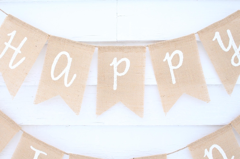 May include: A burlap banner with the word "HAPPY" spelled out in white letters. The banner is made of individual pennants strung together with twine. The background is a white wooden surface.