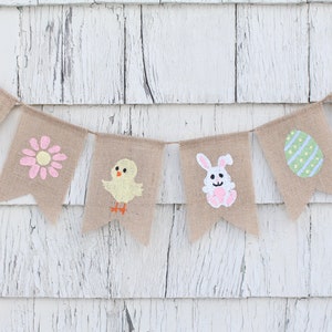 K&ouml;nnte beinhalten: Ein Burlap-Banner mit bemalten Ostern-Motiven. Das Banner zeigt ein K&uuml;ken, einen Hasen, eine Blume, ein Ei und zwei L&auml;mmer. Das Banner ist an einer wei&szlig;en Holzwand befestigt.