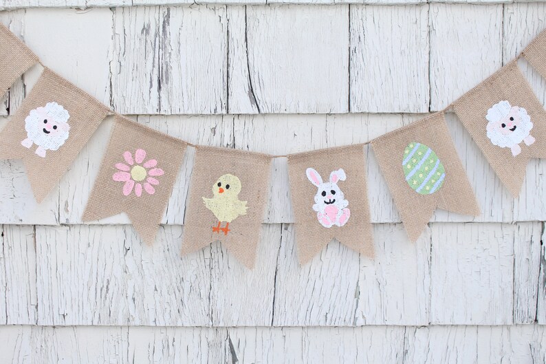 K&ouml;nnte beinhalten: Ein Burlap-Banner mit bemalten Ostern-Motiven. Das Banner zeigt ein K&uuml;ken, einen Hasen, eine Blume, ein Ei und zwei L&auml;mmer. Das Banner ist an einer wei&szlig;en Holzwand befestigt.