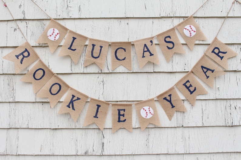 Pu&ograve; includere: Uno striscione in juta con il testo "Luca's Rookie Year" in lettere blu scuro. Lo striscione &egrave; decorato con palline da baseball ed &egrave; appeso a un muro di legno bianco.