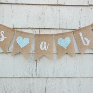 May include: A burlap banner with the words "It's a boy" in white paint. The banner has blue hearts between each word.