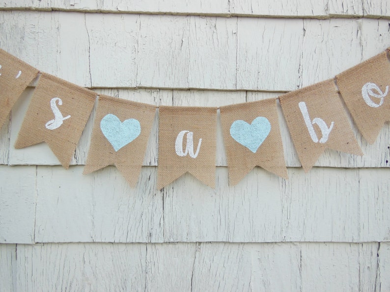 May include: A burlap banner with the words "It's a boy" in white paint. The banner has blue hearts between each word.