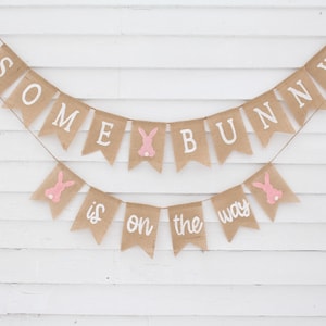 May include: A burlap banner with the text "Some Bunny is on the way" in white lettering. The banner is decorated with pink bunny silhouettes.