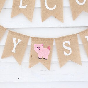 Pode incluir: Uma faixa de juta com letras brancas que dizem "WELCOME BABY". Um desenho de um porco rosa est&aacute; em uma das bandeiras. A faixa est&aacute; pendurada em um fundo de madeira branca.