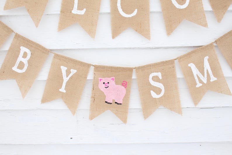 Pode incluir: Uma faixa de juta com letras brancas que dizem "WELCOME BABY". Um desenho de um porco rosa est&aacute; em uma das bandeiras. A faixa est&aacute; pendurada em um fundo de madeira branca.