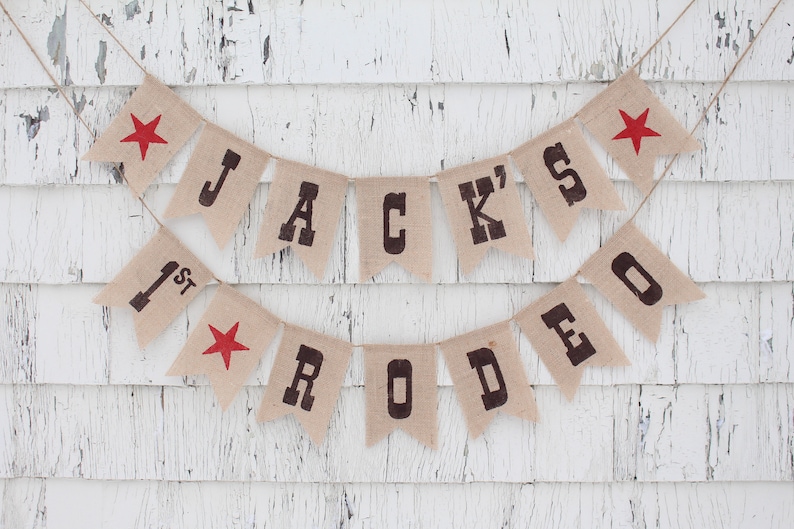 Pode incluir: Uma faixa de serapilheira com o texto "JACK'S 1ST RODEO" em letras castanhas escuras. Estrelas vermelhas decoram as bandeirinhas finais. A faixa &eacute; suspensa por barbante contra um fundo de madeira branca desgastada.