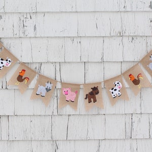 May include: A burlap banner with farm animal illustrations. The banner features a cow, pig, goat, rooster, and horse. The banner is strung on a white wooden wall.