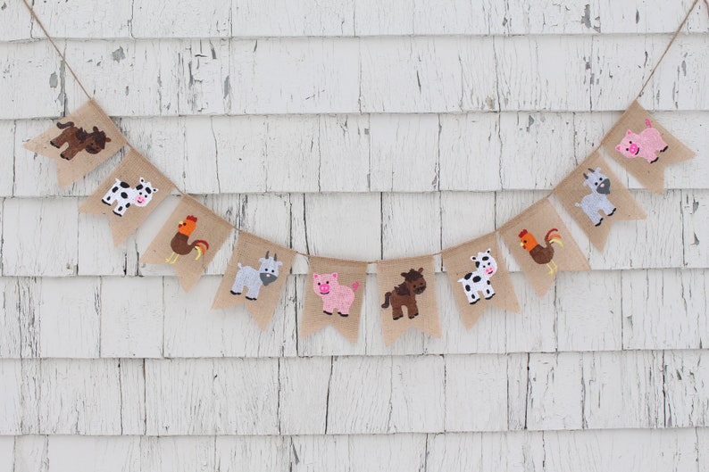 May include: A burlap banner with farm animal illustrations. The banner features a cow, pig, goat, rooster, and horse. The banner is strung on a white wooden wall.
