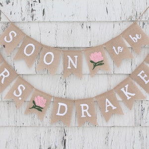May include: A burlap banner with the text "Mrs. Soon to be Drakes" in white letters. The banner is decorated with pink flowers.