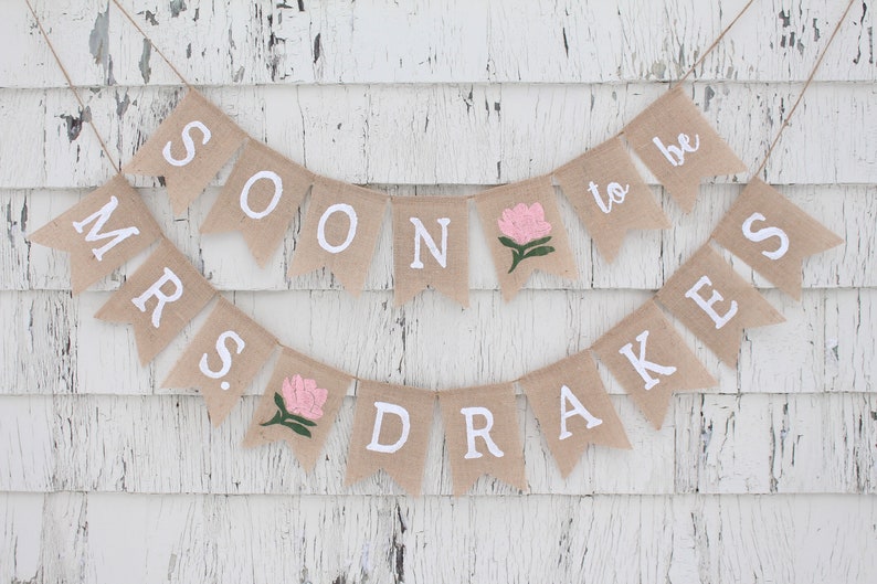 May include: A burlap banner with the text "Mrs. Soon to be Drakes" in white letters. The banner is decorated with pink flowers.