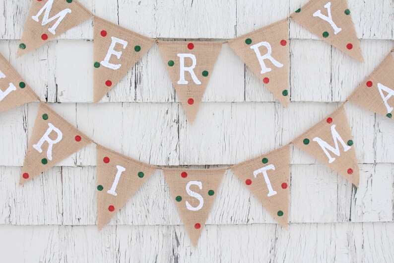 May include: A festive burlap banner spelling "MERRY CHRISTMAS" in white capital letters. Each triangular flag is adorned with red and green dots. The banner is set against a white, weathered wooden backdrop.