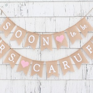 May include: A burlap banner with the text "Mr. & Mrs. Soon to be Carufe" in white letters. The banner has pink hearts between the words.