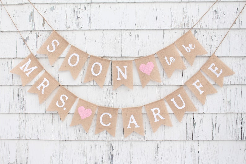 May include: A burlap banner with the text "Mr. & Mrs. Soon to be Carufe" in white letters. The banner has pink hearts between the words.