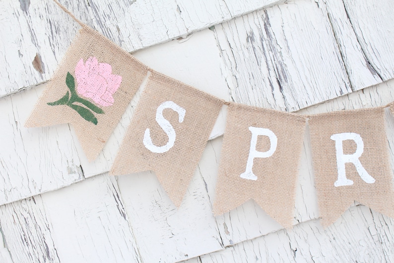 May include: A burlap banner with the word "SPRING" spelled out in white paint. The first flag has a pink flower with green leaves.