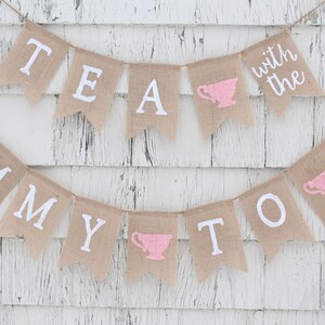 May include: A burlap banner with the text "Tea with the Mommy to Be" in white paint. The banner has pink teacup silhouettes on each end.