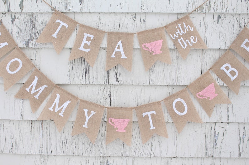 May include: A burlap banner with the text "Tea with the Mommy to Be" in white paint. The banner has pink teacup silhouettes on each end.