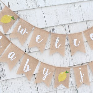 May include: A burlap banner with the words "Welcome Baby" in white paint. The banner is decorated with small yellow lemons.