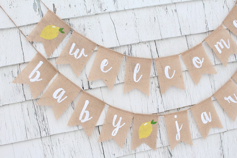 May include: A burlap banner with the words "Welcome Baby" in white paint. The banner is decorated with small yellow lemons.