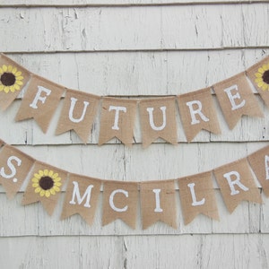 May include: A burlap banner with the words "Future Mrs. Mcllrath" and sunflowers. The banner is strung on a white wooden wall.