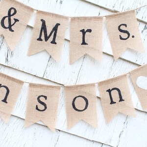 K&ouml;nnte beinhalten: Ein Banner aus Jute mit den Worten "Mr. & Mrs. Johnson" in schwarzer Schrift und einem wei&szlig;en Herz auf der letzten Flagge. Das Banner ist an einer wei&szlig;en Holzwand befestigt.